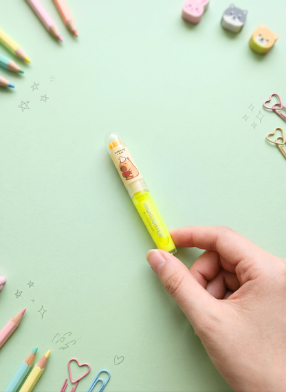 Hand holding a yellow highlighter with cartoon character on a light green background with stationery items.