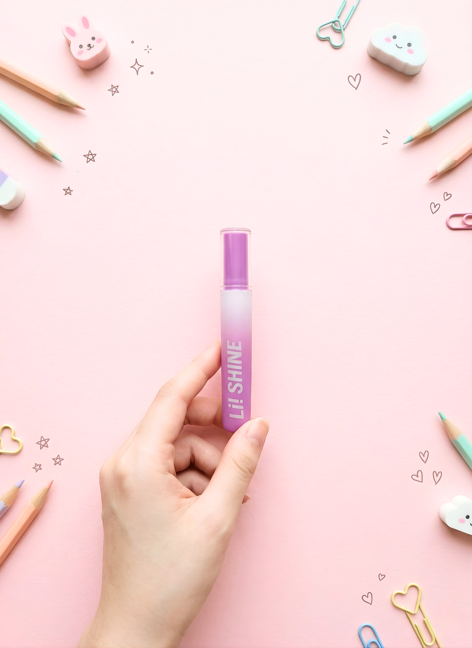Hand holding a purple lip gloss with decorative stationery items on a pink background