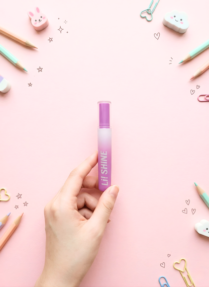 Hand holding a purple lip gloss with decorative stationery items on a pink background