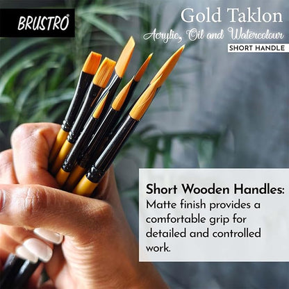 BRUSTRO Gold Taklon paintbrushes with short wooden handles held in a hand, against a blurred indoor background.