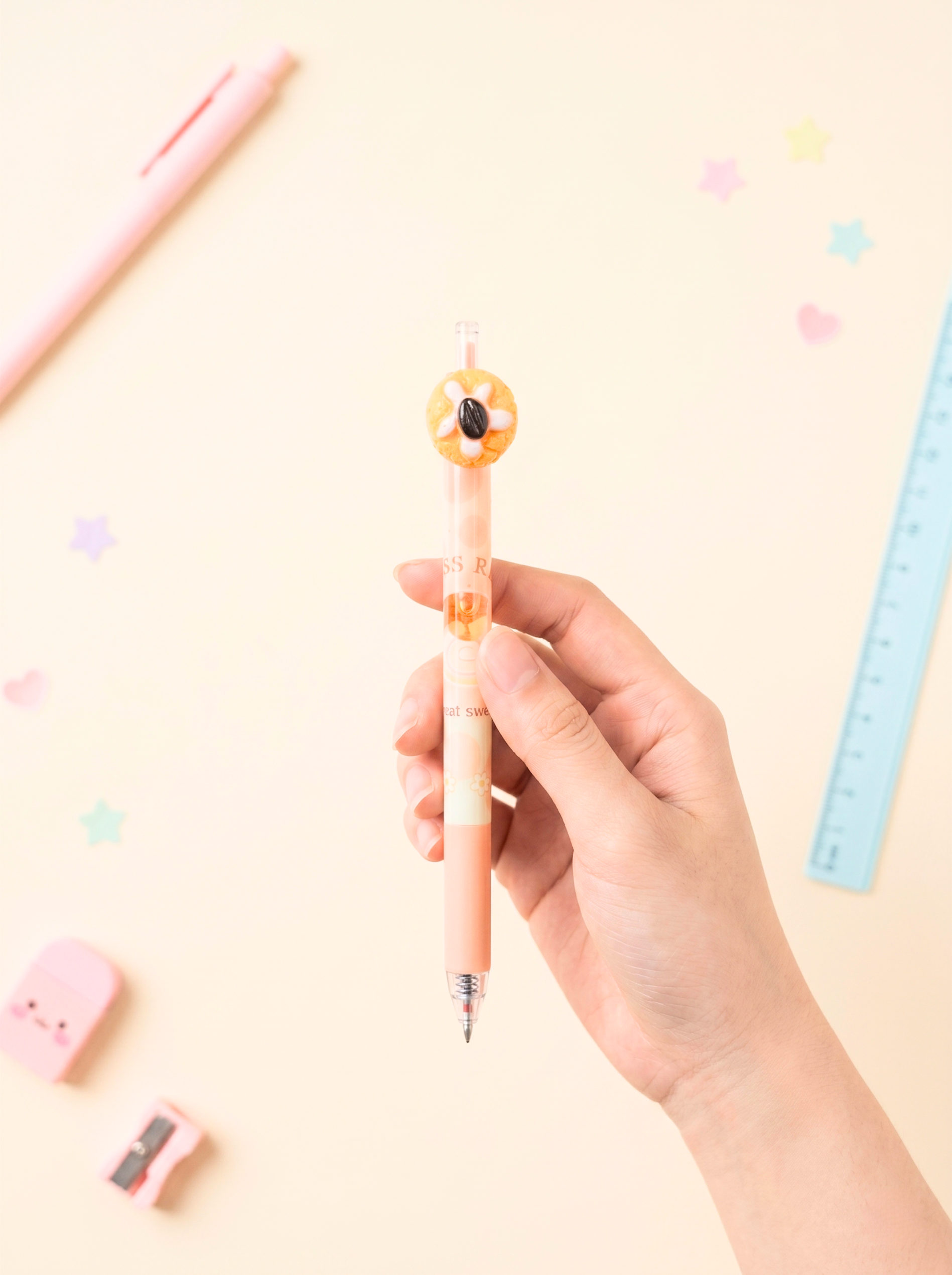 Hand holding a pen with a donut design on a light pink background with stationery items.