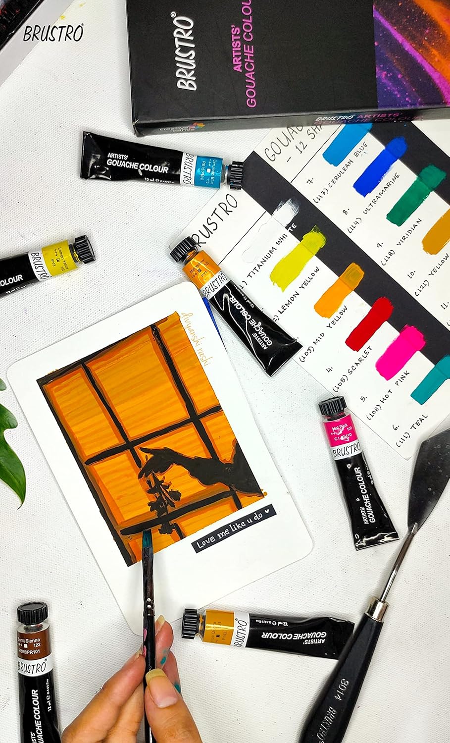 Art supplies including tubes of paint and a painting on paper with a hand holding a brush.