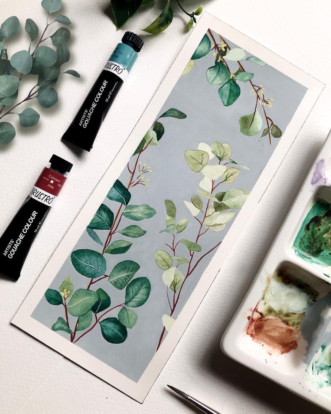 Art supplies including paint tubes and a watercolor painting of leaves on a white surface.