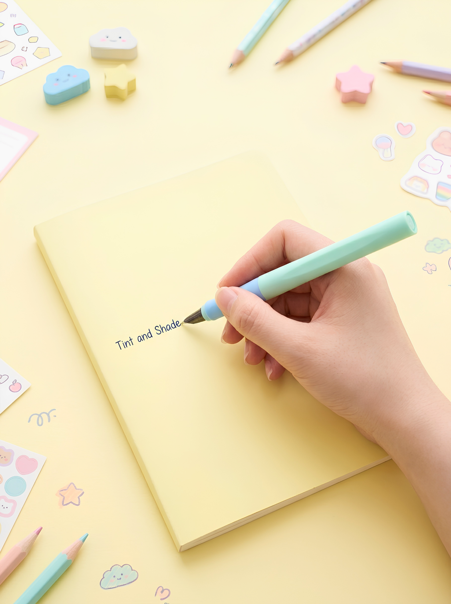 Hand holding a light blue pen writing on a yellow notepad with colorful stationery items around.