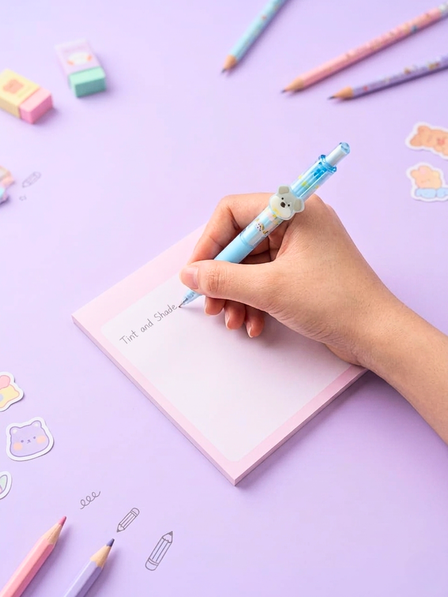 Hand holding a pen with a bear eraser on a notebook with stickers on a purple background