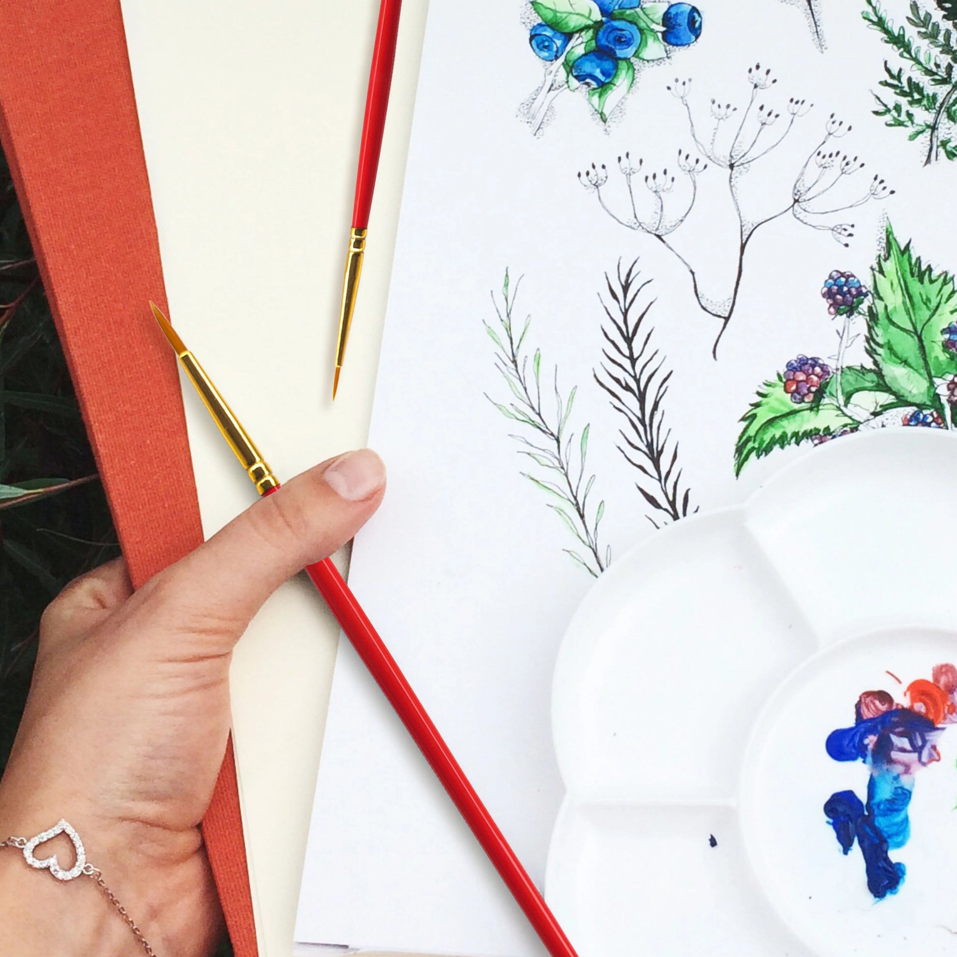 Hand holding a paintbrush with watercolor painting of plants and flowers on paper