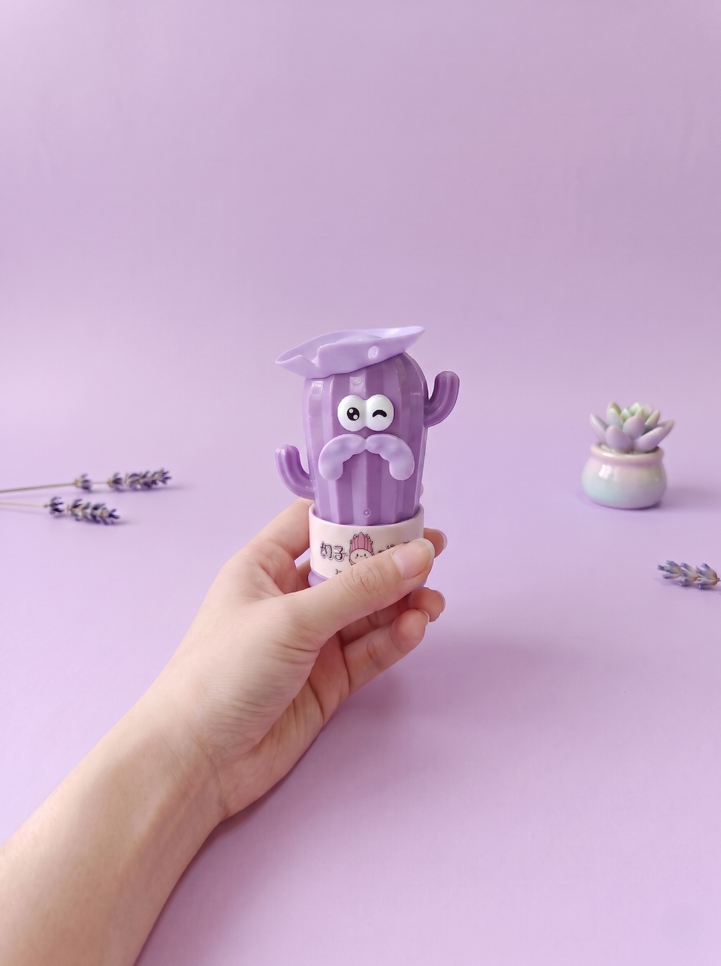 Hand holding a purple cartoon character container against a light purple background with small plants.