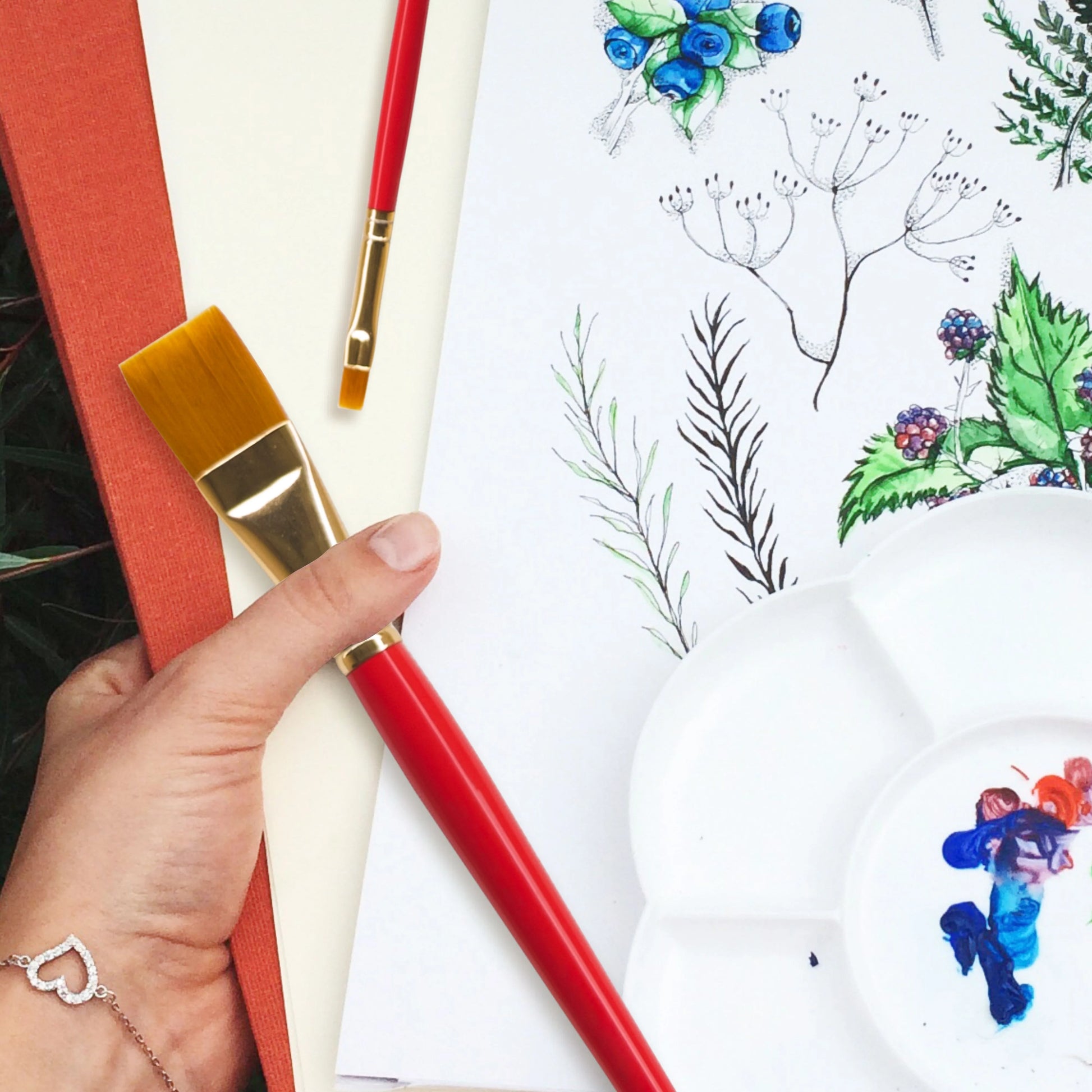 Artistic setup with a hand holding a red paintbrush, watercolor paper, and a palette with floral designs.
