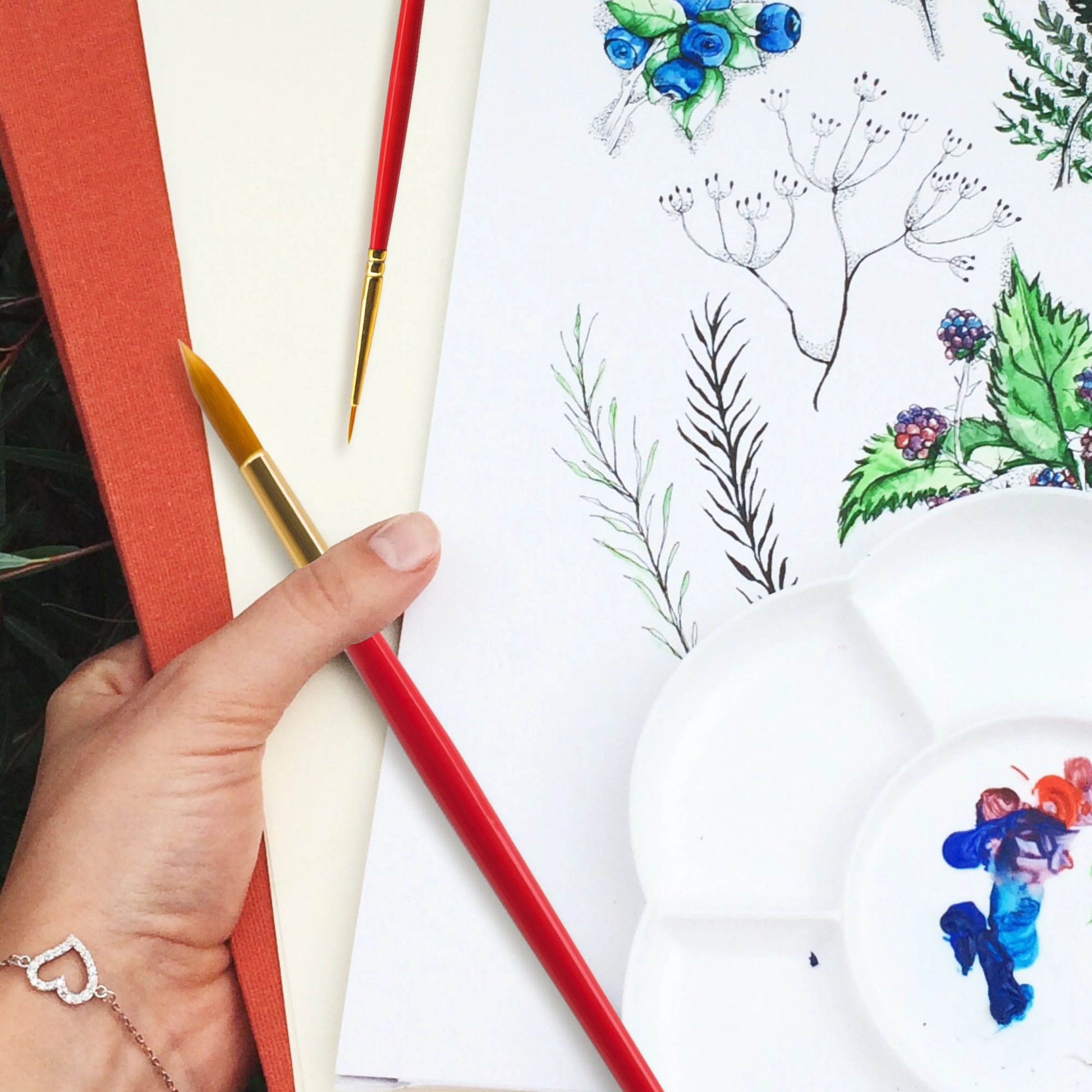 Hand holding a red pencil near a drawing of plants and flowers on a white surface.