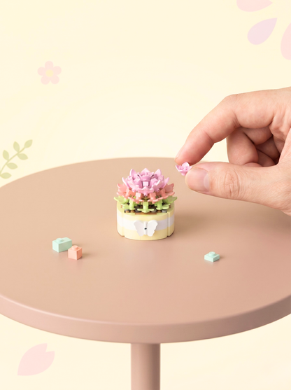 Small decorative cake with pink flowers on a round table, with a hand reaching towards it.
