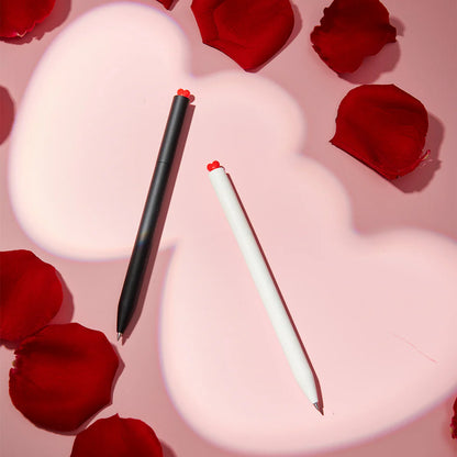 Two pens, one black and one white, on a pink heart-shaped background with red rose petals.