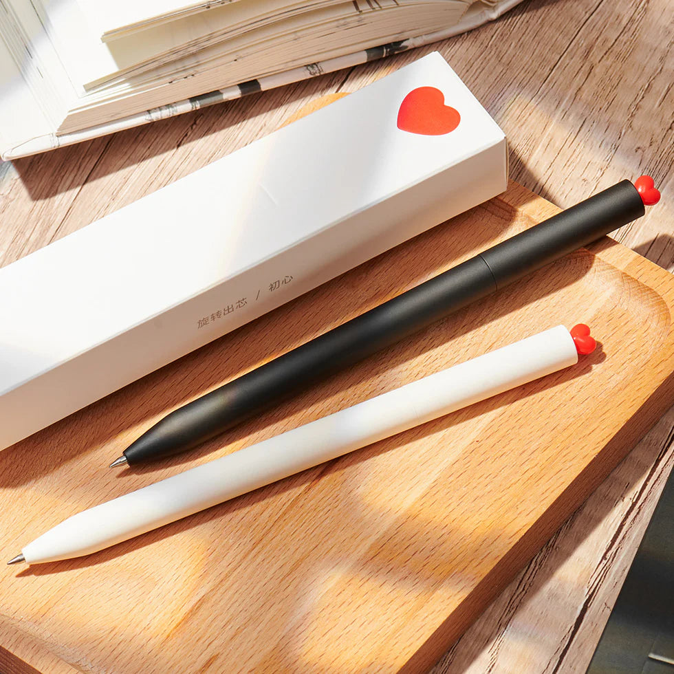 Two pens, one black and one white, on a wooden surface with a heart-shaped object in the background.