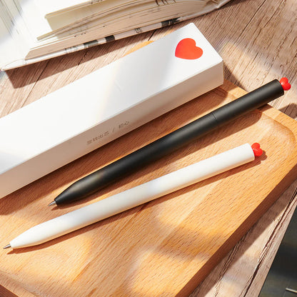 Two pens, one black and one white, on a wooden surface with a heart-shaped object in the background.