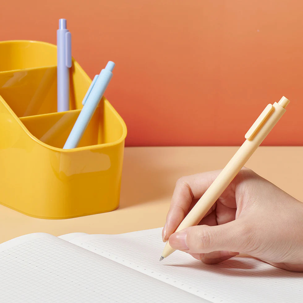 Hand holding a beige pen over a notebook with a yellow pen holder containing pens in the background.