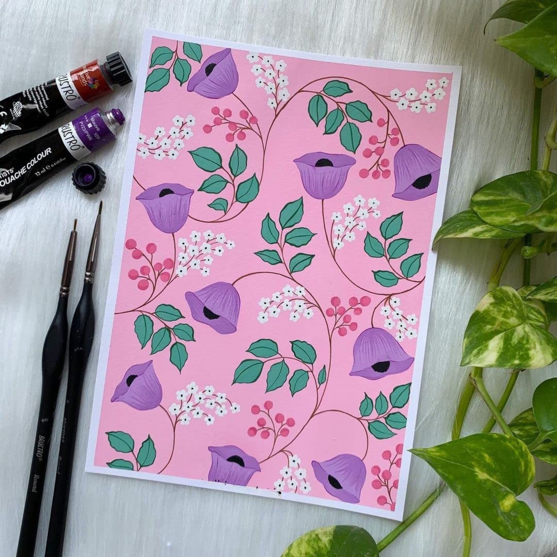 Card with purple floral design on pink background, surrounded by art supplies and a plant.
