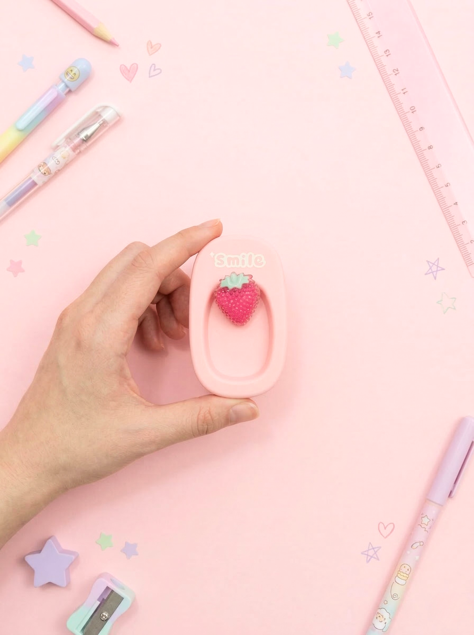 Hand holding a pink strawberry-shaped eraser with stationery items on a pink background