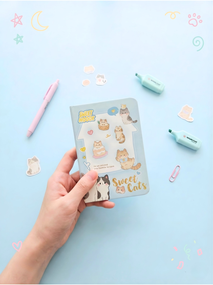 Hand holding a pack of 'Sweet Cats' stickers on a light blue background with stationery items.