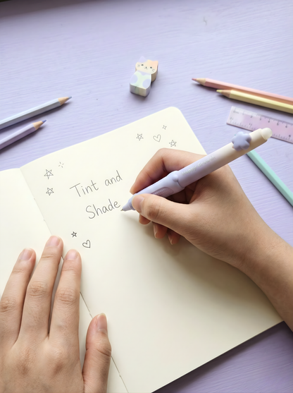 Hand writing 'Tint and Shade' on a notebook with stationery items on a purple surface.