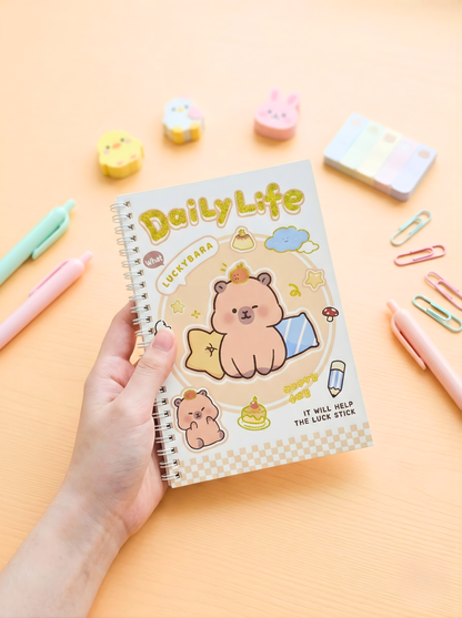 Notebook titled 'Daily Life' with a bear illustration held by a hand on a pastel background with stationery items.