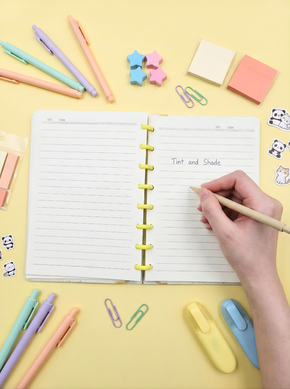 Hand holding a pencil over an open notebook with stationery items on a yellow background