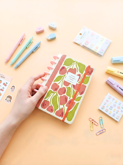 Notebook with cherry design held by a hand on a yellow background with stationery items.