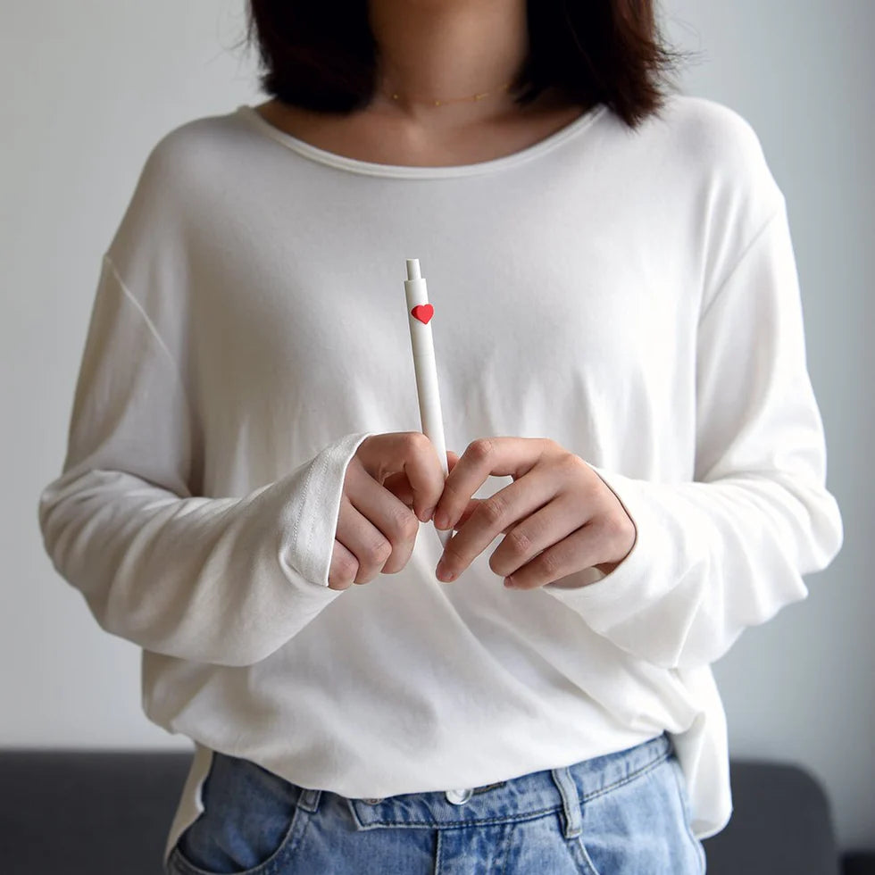 Person wearing a white long-sleeve shirt holding a pen with a red heart on a plain background