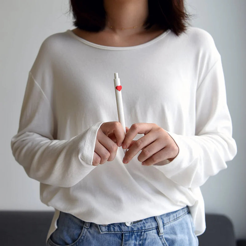 Person wearing a white long-sleeve shirt holding a pen with a red heart on a plain background