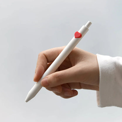 Hand holding a white pen with a red heart-shaped clip on a light gray background