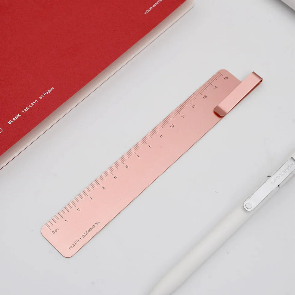 Rose gold ruler on a white surface with a red notebook and white pen.